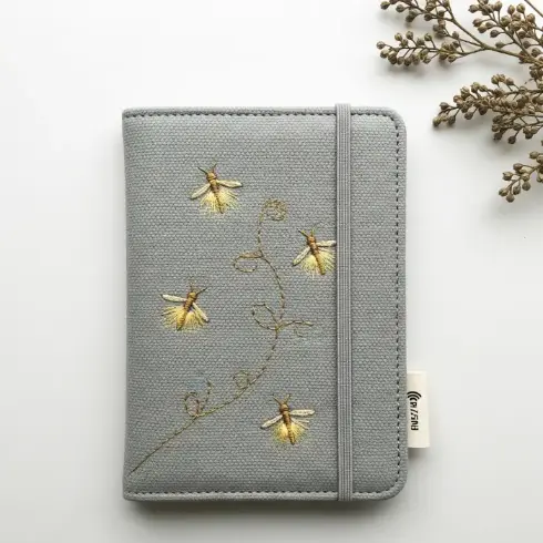 A soft gray recycled canvas passport cover with glowing firefly embroidery in gold thread.
