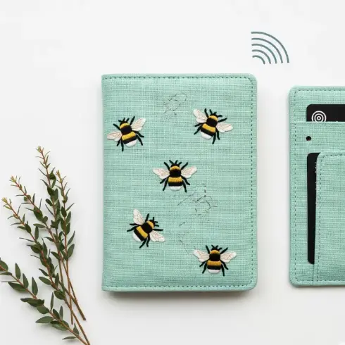 

: A mint green linen passport cover with hand-stitched bumblebees in gold and black thread.
