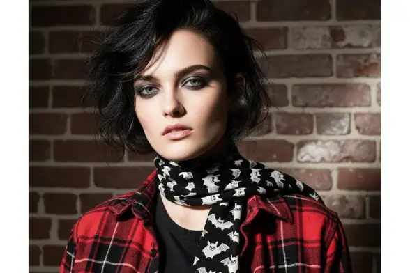 A model in a plaid flannel and bat-print scarf holds a pumpkin in a patch, ready for a spooky fall adventure.