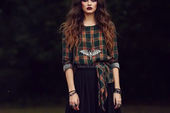 A model wears a patchwork skirt and plaid shirt with a bat-wing pendant, standing by a rustic campfire, capturing a bewitched boho fall look.