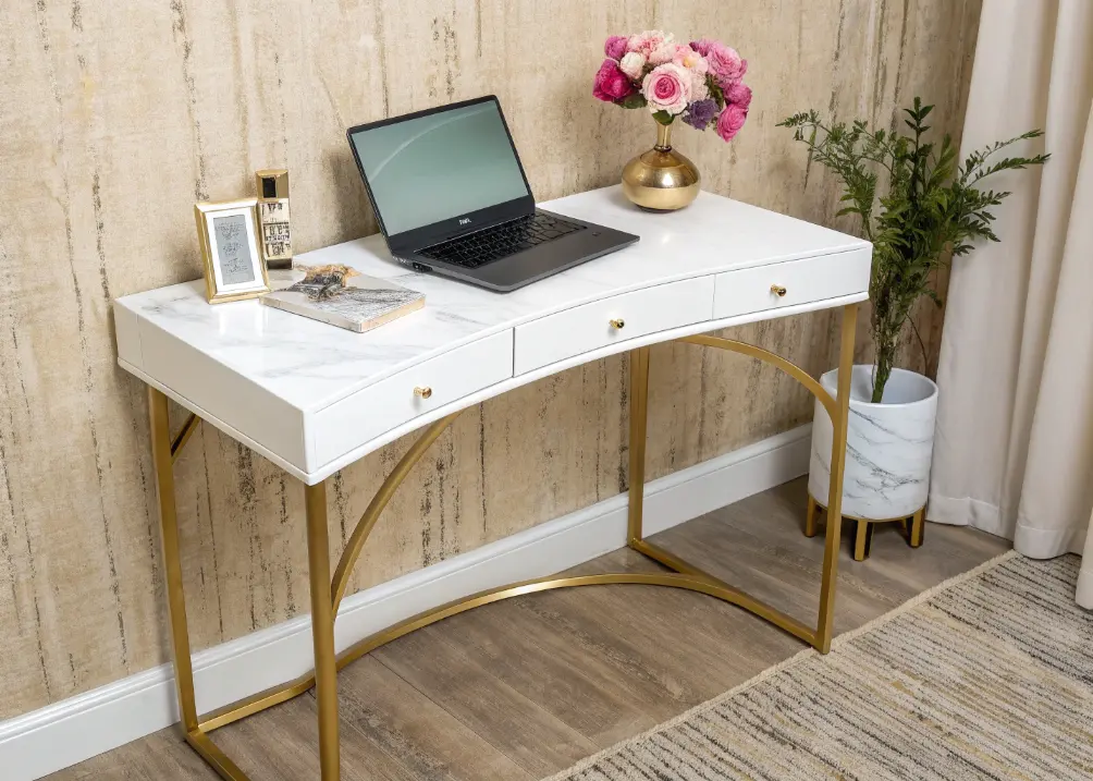 A curved white desk with gold legs, topped with a laptop, a small vase of peonies, and a marble organizer, against a textured beige wall.