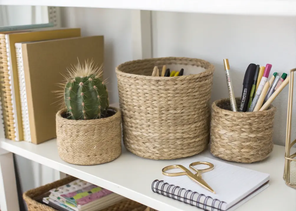 A shelf with woven seagrass baskets holding office supplies, paired with a small cactus and a gold-accented notebook.