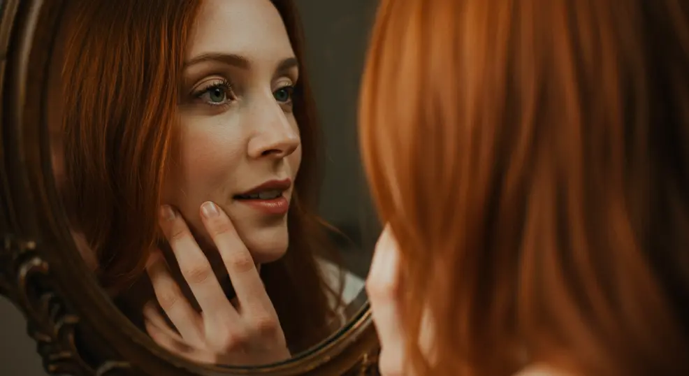 Close-up of a woman looking at her reflection, gently touching her cheek.